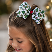 Close-up of a girl wearing a large white Christmas hair bow with green Christmas trees, Santa-hat mascot designs, and a red center wrap, with a decorated Christmas tree in the background.