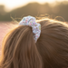 A close-up, side-profile lifestyle shot of a person with wavy brown hair tied into a ponytail using the white Buc-ee’s Easter scrunchie. The scrunchie's festive pattern of pastel eggs, butterflies, and the Buc-ee’s beaver mascot is visible against the hair. The background is a soft-focus field of pink flowers at sunset, creating a warm, golden-hour glow that highlights the texture of the hair and the details of the scrunchie.