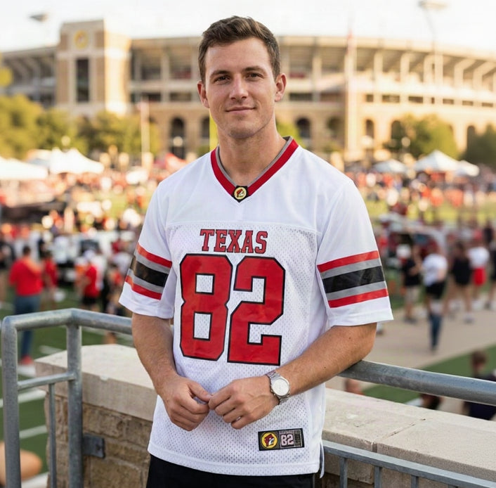 A portrait photograph of a man in a Buc-ee's Football Jersey, a custom white, red, and grey 'Texas 82' football-style jersey, standing and leaning on a metal railing with a large, blurred open-air stadium background filled with people.