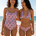 Two smiling women stand arm-in-arm on a sunny beach, wearing coordinating Buc-ee's Pink and Blue Leopard swimwear. One woman wears a one-piece swimsuit with side-ties at the hips, while the other wears a triangle bikini top with matching side-tie bottoms. The background features a turquoise ocean and white sand under a clear sky.
