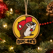 Close-up lifestyle image of a wooden Buc-ee’s Christmas ornament hanging on a decorated Christmas tree, featuring a red-capped beaver on a yellow background with Buc-ee’s lettering and warm white holiday lights.