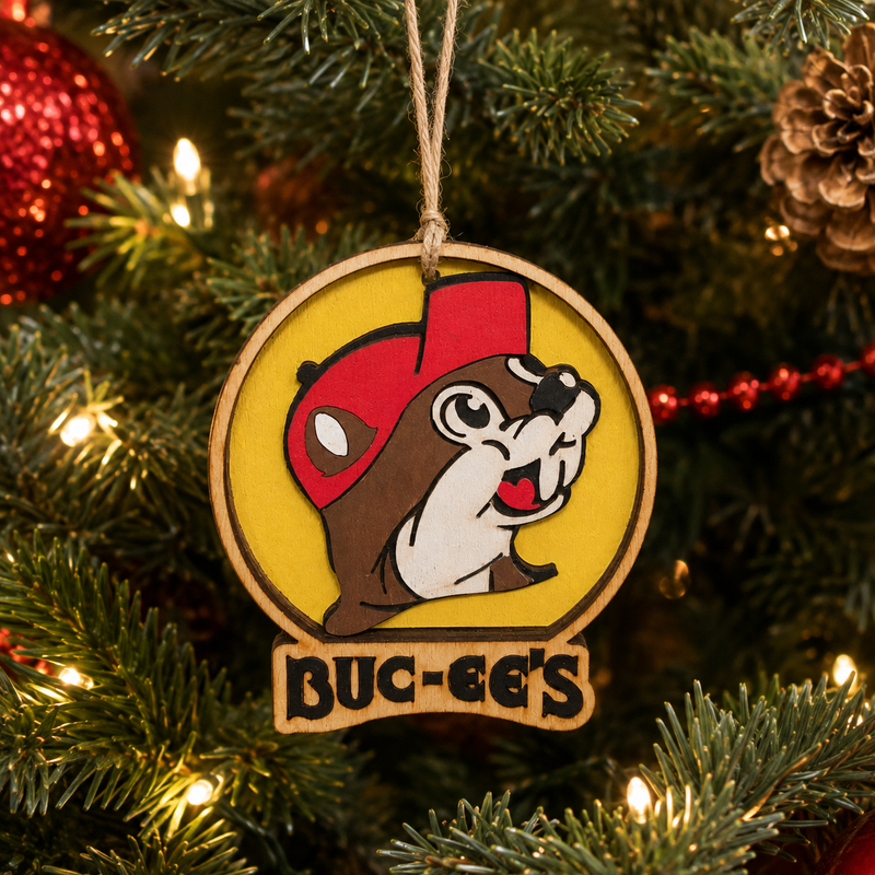 Close-up lifestyle image of a wooden Buc-ee’s Christmas ornament hanging on a decorated Christmas tree, featuring a red-capped beaver on a yellow background with Buc-ee’s lettering and warm white holiday lights.