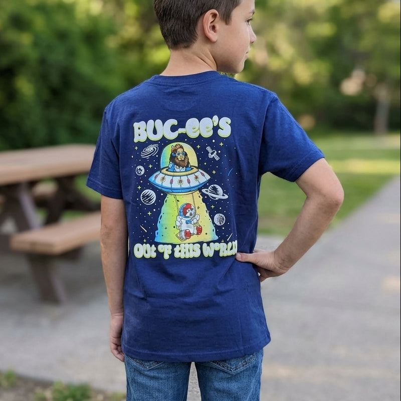 A young boy outdoors wearing the Buc-ee's dark navy blue youth t-shirt, showing the full-color back graphic of Sasquatch in a UFO beaming down astronaut Buc-ee with the words "Out Of This World."