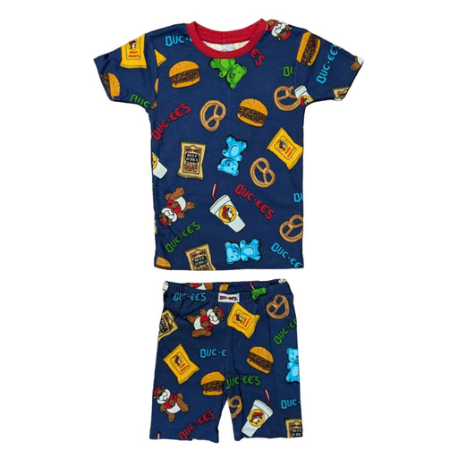 A flat-lay product photograph of a Buc-ee's youth two-piece pajama set on a clean white background. The set includes a short-sleeve top and matching shorts in dark blue, featuring an all-over cartoon print of Buc-ee the Beaver, brisket sandwiches, pretzels, gummy bears, Beaver Nuggets bags, and beef jerky. The shirt has a contrasting bright red crewneck collar.