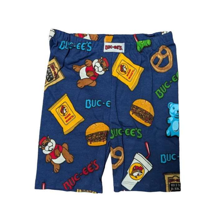 A product photograph of a pair of Buc-ee's youth pajama shorts centered on a clean white background. The dark blue shorts feature a flexible elastic waistband and an all-over cartoon print of Buc-ee the Beaver alongside iconic road trip treats, including bags of Beaver Nuggets, beef jerky, sliced brisket sandwiches, gummy bears, pretzels, and fountain drinks.