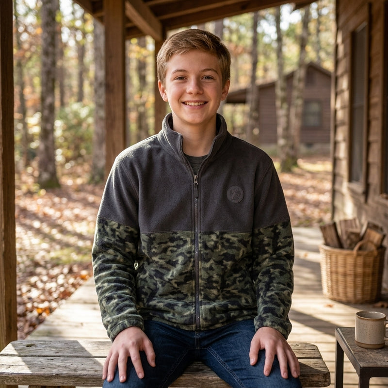 Young boy smiling while sitting on a porch bench, wearing a Buc-ee's Camo Colorblock Fleece Jacket, half-solid charcoal and half-camouflage fleece jacket.