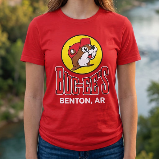 A smiling woman stands outdoors in front of a river and forest canyon, wearing a bright red graphic T-shirt with a yellow circular bug-eat logo and the text 'BUG-EATS BENTON, AR'.