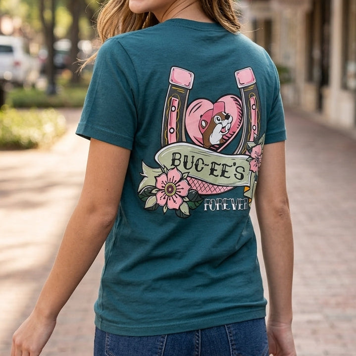 A woman wearing a teal graphic t-shirt with a 'Bueee's Bachelorette' horseshoe and nail design, walking down a sunny brick sidewalk.