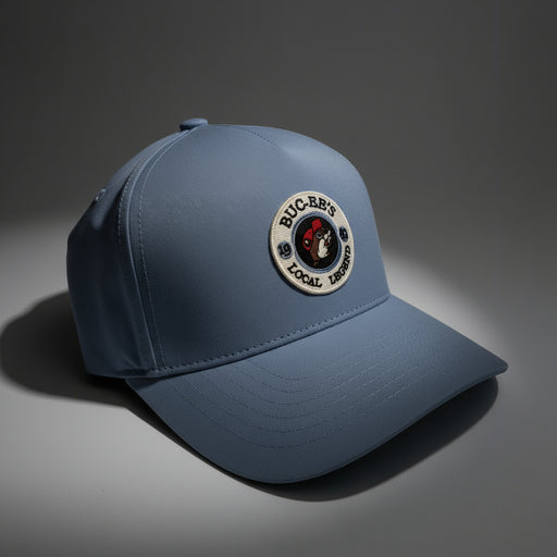 Close-up of a blue Buc-ee’s “Local Legend” baseball cap with a round embroidered patch featuring the Buc-ee’s logo. The hat rests under soft lighting on a neutral background, showing off its smooth fabric texture and structured shape.