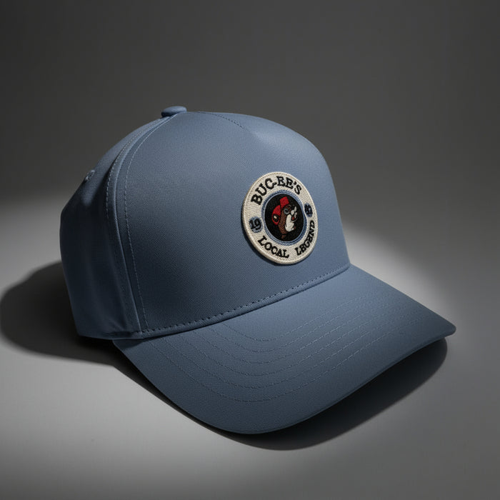 Close-up of a blue Buc-ee’s “Local Legend” baseball cap with a round embroidered patch featuring the Buc-ee’s logo. The hat rests under soft lighting on a neutral background, showing off its smooth fabric texture and structured shape.