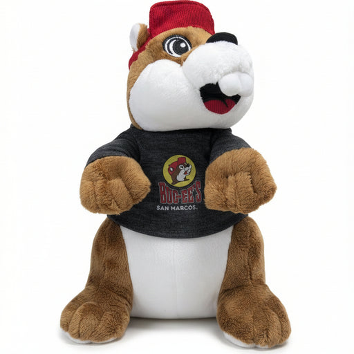 A San Marcos mascot plush toy sitting upright, wearing a red cap and a black t-shirt with the San Marcos logo.