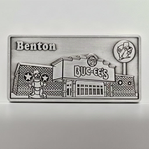 A rectangular silver-toned metal plaque featuring an engraved illustration of a Buc-ee's store front with the word 'Benton' at the top left and a circular logo at top right, shown on a plain white background.