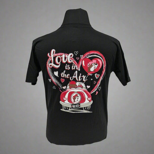 A black T-shirt featuring a colorful Valentine's Day-themed design on the back. The graphic includes two characters in a red car with a Buc-ee's logo on the license plate, surrounded by a large pink heart and smaller white hearts. The text reads "Love is in the Air" in a playful script font.




