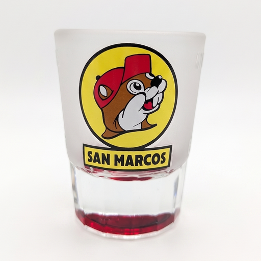 A frosted glass shot glass featuring the San Marcos mascot logo, a bulldog wearing a red cap inside a yellow circle, with 'SAN MARCOS' printed below. The bottom of the glass has a red tint, and it is photographed on a clean white background.
