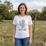 Woman wearing a white t-shirt with a colorful cactus design and 'Happy Easter' text, standing in a field with wildflowers.
