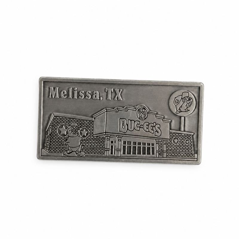 A rectangular pewter metal magnet showing an embossed 3D relief of the Buc-ee's Melissa travel center building, complete with the beaver statue out front, the Buc-ee's sign, a lollipop road sign in the upper right, and the word "Melissa" in the upper left corner.