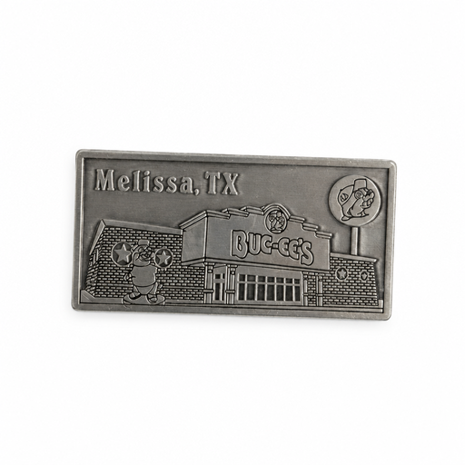 A rectangular pewter metal magnet showing an embossed 3D relief of the Buc-ee's Melissa travel center building, complete with the beaver statue out front, the Buc-ee's sign, a lollipop road sign in the upper right, and the word "Melissa" in the upper left corner.