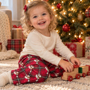 A young toddler girl with blonde curly hair sits on a beige carpet in front of a decorated Christmas tree, wearing a cream long-sleeve shirt and red plaid pajama pants with a snowman pattern. She is smiling and interacting with a small wooden toy truck.