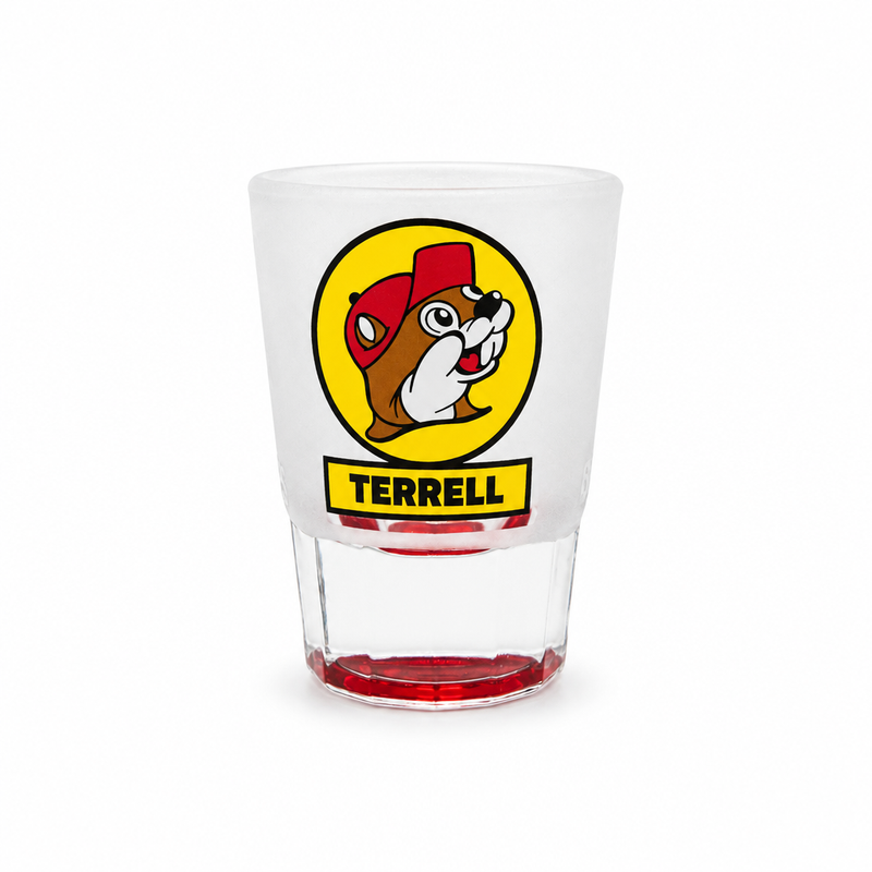 A clear frosted shot glass featuring the yellow circular Buc-ee's beaver logo with a yellow banner underneath reading "TERRELL" in black text, set against a solid white background. The glass features a thick clear base tinted red at the very bottom.
